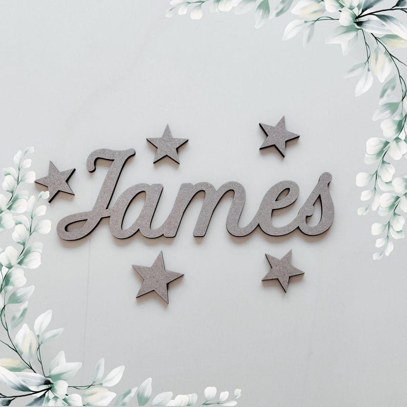 May include: Wooden name sign with the name "James" in a cursive font, accompanied by several star-shaped cutouts. The sign and stars are a muted gray color, set against a light gray background with decorative floral accents.