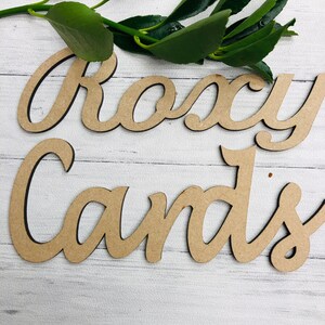 May include: Two wooden cutouts spelling "Roxy Cards" in a cursive font. The letters are a light brown color with a dark brown outline. Green leaves are in the background, and the letters are on a white wooden surface.