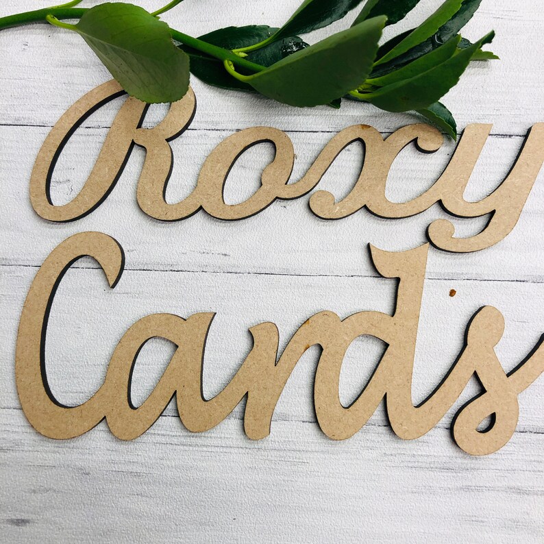 May include: Two wooden cutouts spelling "Roxy Cards" in a cursive font. The letters are a light brown color with a dark brown outline. Green leaves are in the background, and the letters are on a white wooden surface.