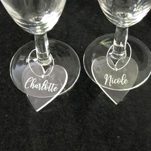 May include: Two clear glass champagne flutes with heart-shaped acrylic charms attached. The charms are engraved with the names "Charlotte" and "Nicole".