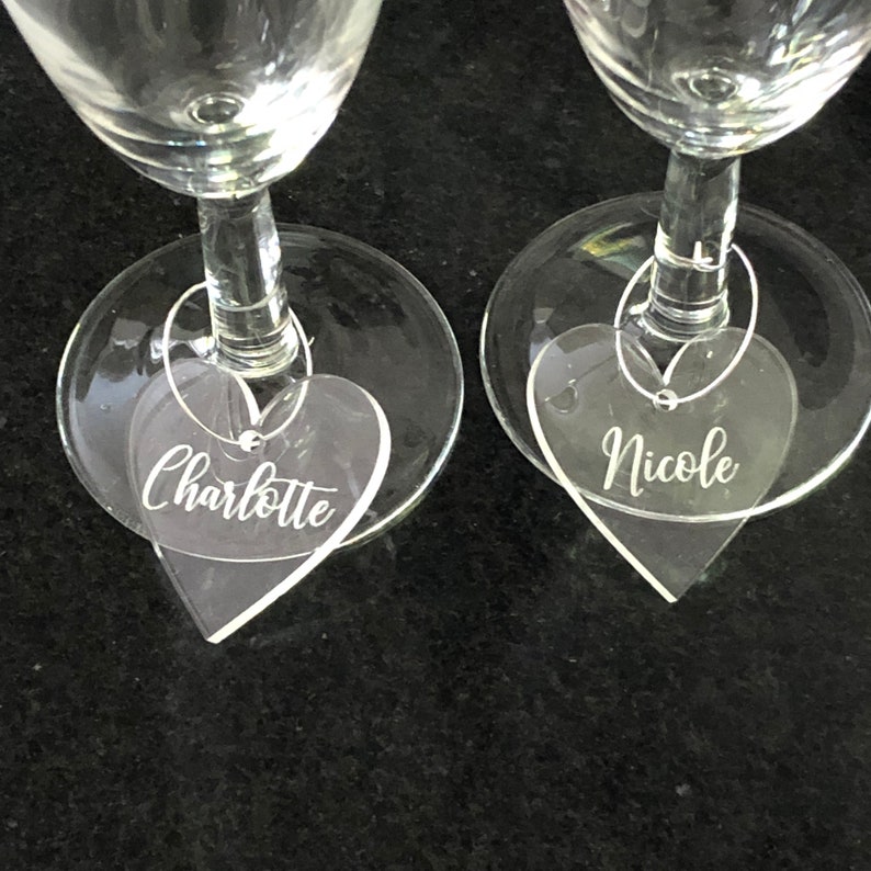 May include: Two clear glass champagne flutes with heart-shaped acrylic charms attached. The charms are engraved with the names "Charlotte" and "Nicole".