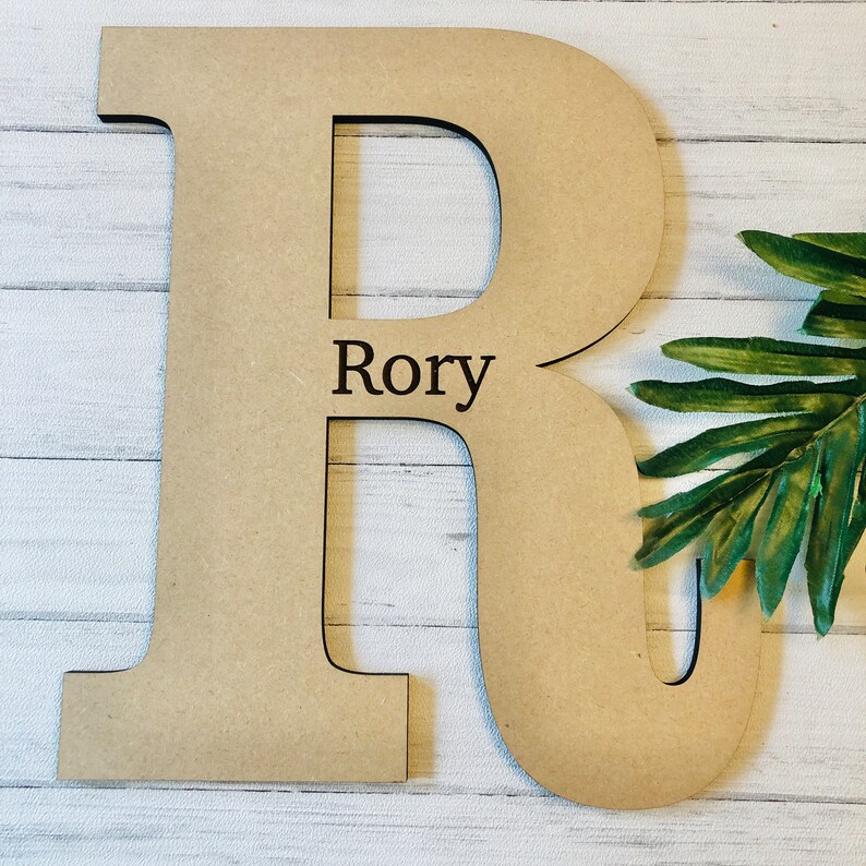 Pode incluir: Uma grande letra R de madeira, pintada de cor castanho claro. A letra &eacute; independente e tem um design recortado. O nome "Rory" est&aacute; gravado na letra.