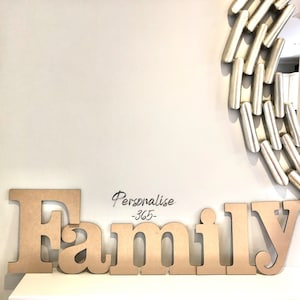 May include: A large, light brown wooden cutout sign spelling the word Family in a serif font. Above the word, the text Personalise-365- is printed in a black script font. A portion of a silver, cylindrical-linked circular wall mirror is visible.
