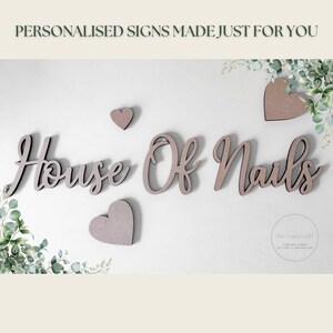 May include: Wooden sign with the text "House Of Nails" in a cursive font, along with three heart-shaped decorations. The sign is light brown and mounted on a white wall, with greenery accents.