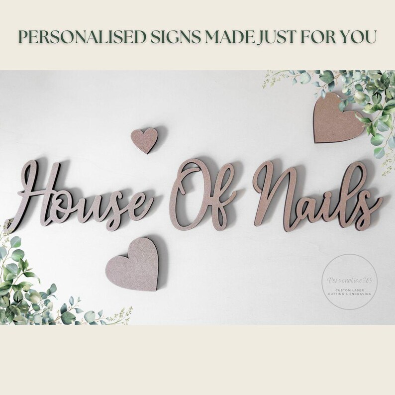 May include: Wooden sign with the text "House Of Nails" in a cursive font, along with three heart-shaped decorations. The sign is light brown and mounted on a white wall, with greenery accents.