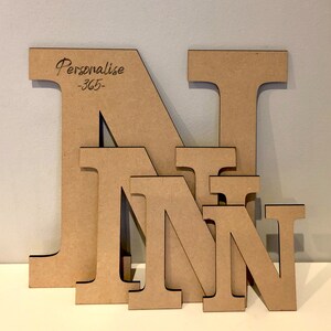 May include: Four light brown wooden letters N of varying sizes stacked in front of each other. The largest letter features the engraved text Personalise -365- in the upper left corner. Each letter has a smooth, flat surface and dark edges.