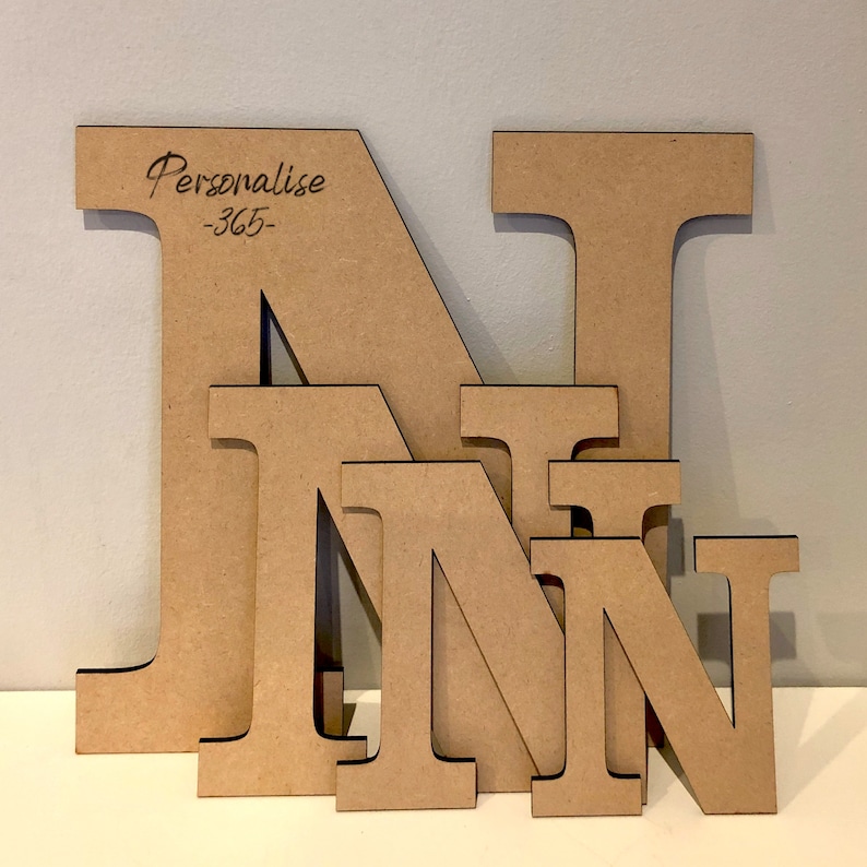 May include: Four light brown wooden letters N of varying sizes stacked in front of each other. The largest letter features the engraved text Personalise -365- in the upper left corner. Each letter has a smooth, flat surface and dark edges.