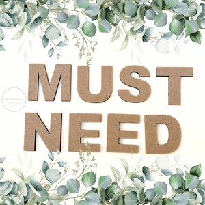 May include: A set of brown, flat, capital letters made of wood spelling out the words MUST NEED. The letters are arranged in two rows against a white background decorated with green and blue eucalyptus leaves at the top and bottom edges.