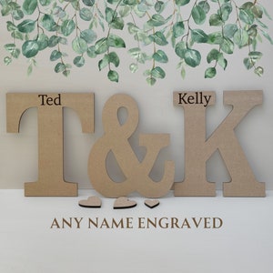 Pode incluir: Letras de madeira que soletram "T & K" com os nomes "Ted" e "Kelly" gravados acima. Tr&ecirc;s pequenos cora&ccedil;&otilde;es de madeira est&atilde;o abaixo das letras. O texto "ANY NAME ENGRAVED" est&aacute; abaixo dos cora&ccedil;&otilde;es.