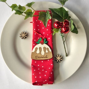 May include: A red napkin with white stars is tied with a wooden Christmas ornament shaped like a pudding. The ornament has the name "David" written on it. The ornament is hanging from a sprig of holly.