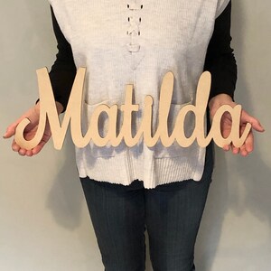 May include: A light-coloured wooden cutout sign featuring the name Matilda in a cursive script font. The letters are connected in a single piece, showcasing a smooth, natural wood finish with a flat surface and clean, laser-cut edges throughout.