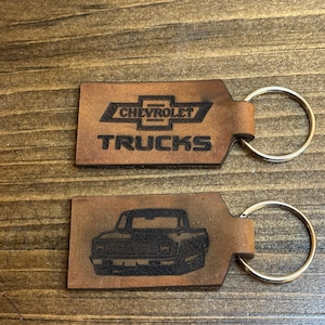 May include: Two brown leather keychains with silver key rings. One keychain features the Chevrolet logo and the text "TRUCKS". The other keychain features a black and white silhouette of a Chevrolet truck.