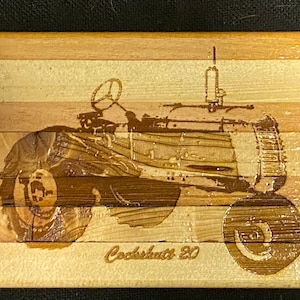 May include: A wooden wall art piece featuring a vintage tractor with the text "Cockshutt 20" engraved on the bottom. The tractor is depicted in a brown silhouette against a light wood background.