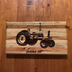 May include: A wood sign with a black and white image of a Cockshutt 550 tractor. The sign is made of multiple pieces of wood, each with a different grain pattern. The text "Cockshutt 550" is burned into the wood below the image.