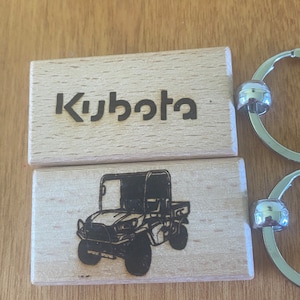 Peut inclure: Deux porte-clés en bois avec un anneau de clé argenté. Le porte-clés du haut porte le mot "Kubota" gravé dessus. Le porte-clés du bas a une image en noir et blanc d'un véhicule utilitaire Kubota.