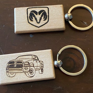 May include: Two wooden keychains with metal key rings. One keychain features a black and white silhouette of a Ram truck. The other keychain features a black and white silhouette of the Ram logo.