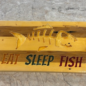 May include: Wooden sign with a fish skeleton design and the words "EAT SLEEP FISH" in colorful letters. The sign is made of light and dark wood planks, creating a rustic look. A decorative item for a fishing enthusiast.