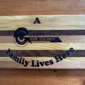 May include: A wooden cutting board with a black and white logo for Cockshutt Farm Equipment. The board is made of different types of wood and has the words "A Family Lives Here" engraved on it.