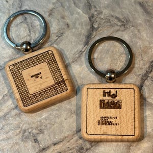 Puede incluir: Dos llaveros de madera con aros plateados. Uno tiene el texto "Pentium" y el otro "Intel i486" grabado en la madera clara. Los llaveros son cuadrados con esquinas redondeadas.