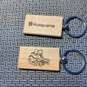 May include: Two wooden keychains with metal rings. One keychain features the Husqvarna logo and the other features a line drawing of a riding lawnmower.