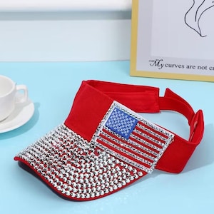 May include: A red visor with a sparkling, silver-colored brim. The visor features a patch with the American flag design, with a blue field of stars and red and silver stripes. The visor has an adjustable strap.