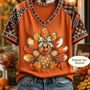 May include: An orange V-neck t-shirt featuring a graphic of a turkey with a polka-dot bow, surrounded by colorful feathers. The sleeves have a patchwork pattern of plaid and leopard print. Text on the shirt reads: Printed, Not Knitted.