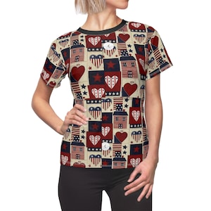 Puede incluir: Camiseta de manga corta con un estampado repetido de corazones rojos, estrellas y casas con diseños de la bandera estadounidense sobre un fondo crema. La camiseta tiene cuello y mangas negros.