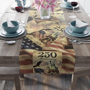 Peut inclure: Une nappe avec un motif patriotique représentant le drapeau américain, la Cloche de la Liberté et la Statue de la Liberté. La nappe porte l'inscription "250e anniversaire" et "1776-2026". La table est dressée avec des assiettes, des bols et des verres à vin.