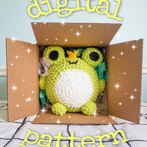 May include: A digital pattern for crocheting a green frog with a white belly and a gold crown. The frog is inside a brown cardboard box with a white and black checkered background.
