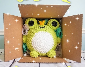 May include: A digital pattern for crocheting a green frog with a white belly and a gold crown. The frog is inside a brown cardboard box with a white and black checkered background.