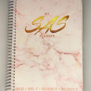 May include: A spiral-bound planner with a pink marble design. The cover features gold lettering that reads "MY SAS PLANNER." Below the title are the words "SEE IT • FEEL IT • BELIEVE IT • RECEIVE IT."