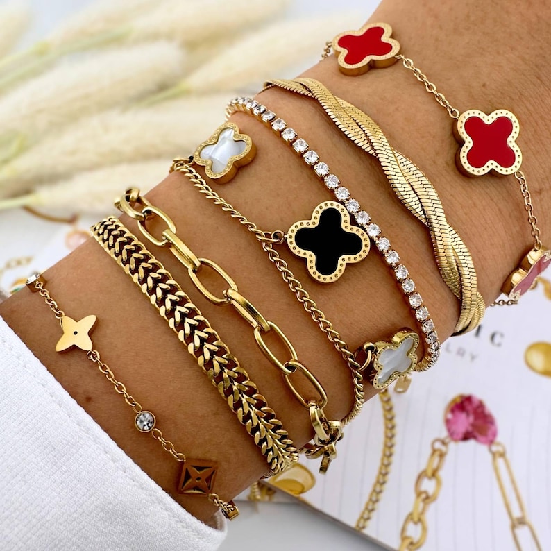 May include: A stack of gold bracelets with different chain styles and charms. The charms include a black clover, a white clover, and a red clover.