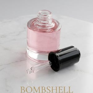 May include: A clear glass bottle with a pink liquid inside, a black cap, and a glass dropper. The word "BOMBSHELL" is printed in gold, with the tagline "- Seductive. Iconic. Elevated." below.