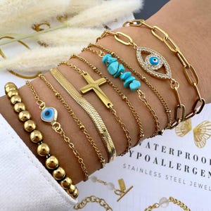 May include: A set of gold-toned bracelets with various charms, including a cross, an evil eye, and turquoise stones. The bracelets are layered on a person's wrist.