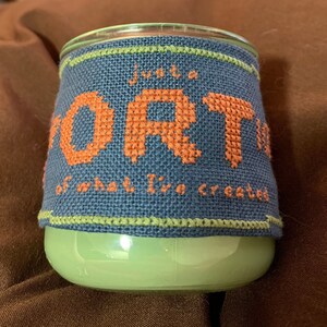 May include: A green glass candle jar with a blue and green fabric cover. The fabric cover has a cross-stitch design that reads "Just a portion of what I've created".