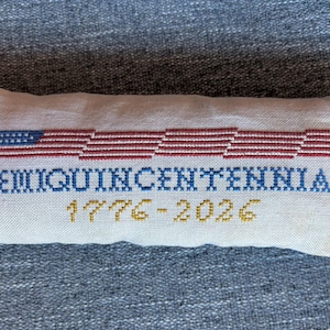 May include: A rectangular, off-white, cross-stitched pillow with a patriotic theme. The design features a stylized American flag in red, white, and blue, along with the word "SEMIQUINCENTENNIAL" and the dates "1776-2026" in gold.