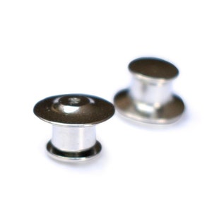 May include: Two small, silver-colored button backs with a flat, circular top and a cylindrical base. The button backs are designed to secure items. The background is plain white, highlighting the metallic finish of the fasteners.