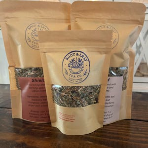 May include: Three brown paper stand-up pouches containing loose-leaf tea, each featuring a circular blue logo with the text Root & Leaf Tea Co. Where Garden Meets The Cup. The pouches have clear windows displaying the dried herbal tea blend inside.