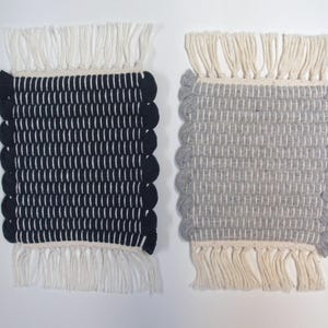 May include: Two rectangular woven coasters. One is navy blue with white vertical lines and a white fringe. The other is gray with white vertical lines and a white fringe. Both have a scalloped edge.