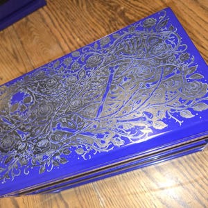 May include: A rectangular, cobalt blue box with a silver floral and skull design. The box has a glossy finish and is resting on a wooden surface. The design includes roses and vines.