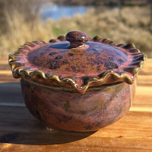 May include: A handcrafted ceramic lidded bowl with a ruffled edge. The bowl and lid are glazed in shades of red, brown, and gold. The lid features a small, round knob. The bowl sits on a wooden surface.