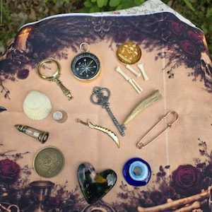 May include: A collection of mystical objects including a compass, magnifying glass, key, heart-shaped glass, and a safety pin, arranged on a patterned cloth with roses and bones. The items vary in color and size, creating a unique display.