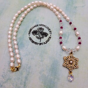 May include: A white pearl necklace with a gold and crystal snowflake pendant. The necklace also features small clear and burgundy beads. The necklace is handmade and has a stamp that reads "Handmade with love by Artistry by Ajanta".