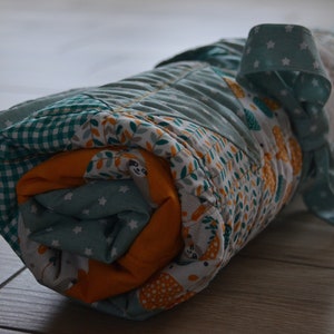 Könnte beinhalten: Eine zusammengerollte Babydecke mit einem Patchwork-Muster in Grün, Weiß und Gelb. Die Decke ist mit einem blau-weißen Band mit weißen Sternen gebunden.