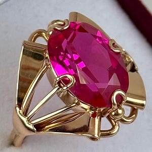 Op de afbeelding: Een gouden ring met een grote, ovaal geslepen, magenta edelsteen. De ring heeft een sierlijk, open ontwerp met gebogen en geometrische elementen. De edelsteen is het middelpunt, gezet in een gedetailleerde gouden zetting.