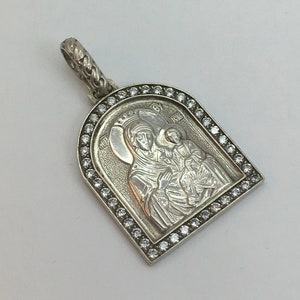 Op de afbeelding: Zilveren hanger met een gegraveerd religieus icoon van een vrouw en kind, omringd door kleine, heldere edelstenen. De hanger heeft een decoratieve beugel voor een ketting.