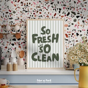 May include: A framed print with the words "So Fresh and So Clean" in dark green lettering. The background of the print has vertical light green stripes. The print is in a light wood frame. The background is a terrazzo pattern.