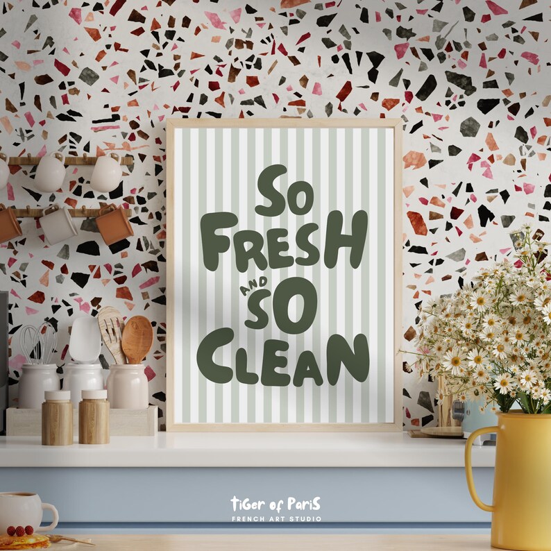 May include: A framed print with the words "So Fresh and So Clean" in dark green lettering. The background of the print has vertical light green stripes. The print is in a light wood frame. The background is a terrazzo pattern.
