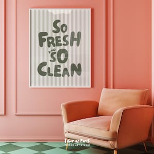 May include: A framed print with the words "So Fresh and So Clean" in a playful font. The background has vertical stripes in shades of gray. The print hangs on a pink wall above a peach-colored armchair.