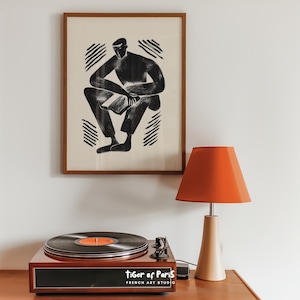 Puede incluir: Una impresión artística en blanco y negro enmarcada de una persona sentada con un libro cuelga sobre un tocadiscos y una lámpara naranja. El tocadiscos es marrón con un disco negro en el plato giratorio. La lámpara tiene una base beige y una pantalla naranja.